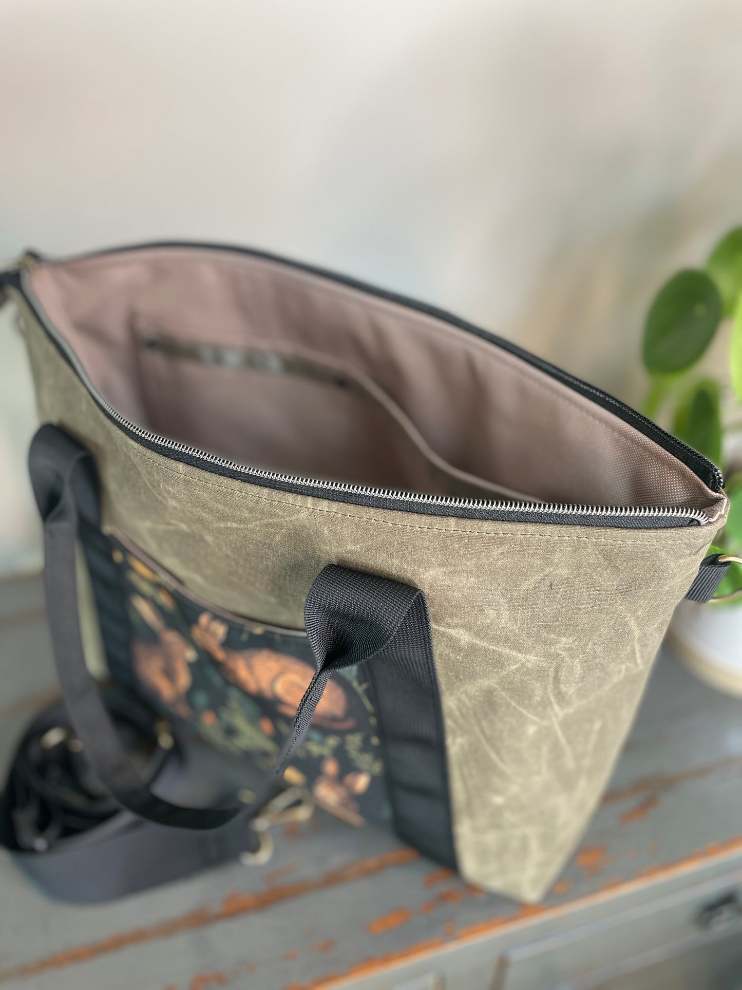 Oxbow crossbody/tote bag in waxed canvas exterior, with black nylon adjustable strap and handles. Two exterior front pockets, multiple inner pocket, fully lined. Olive green waxed canvas, rabbit print on accent front and back pockets.