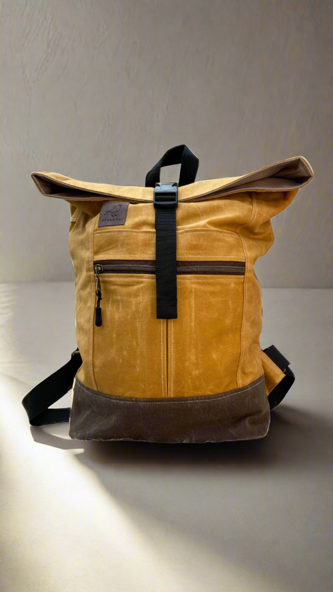 Roll-top backpack in ochre (yellow) waxed canvas and a brown waxed canvas base. Nylon webbing straps, buckle closure on top, hang tab on top. Zippered expandable front pocket. Unisex.