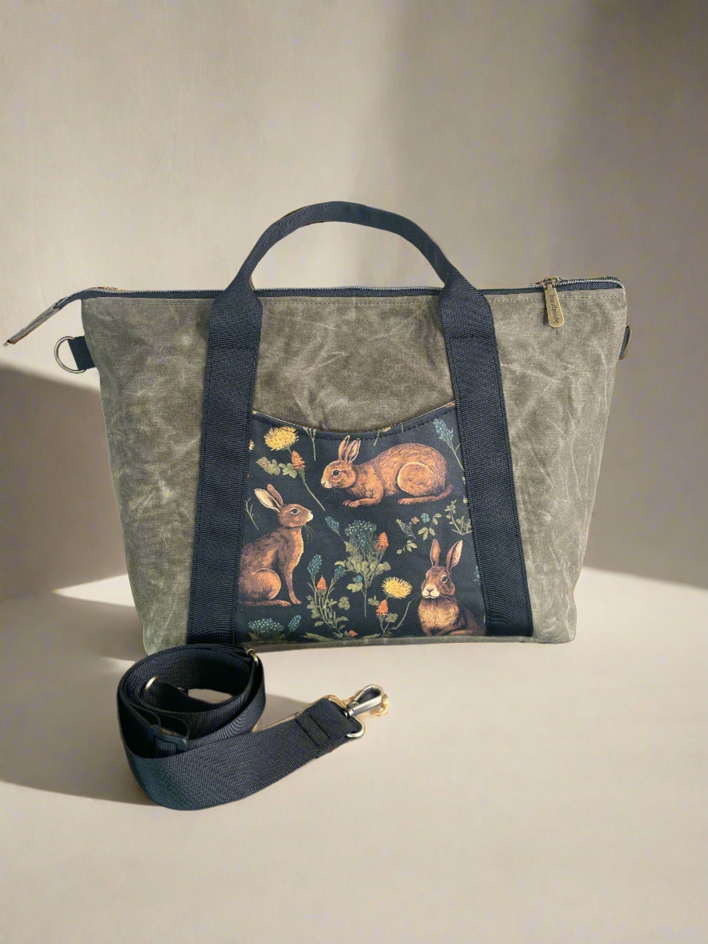 Oxbow crossbody/tote bag in waxed canvas exterior, with black nylon adjustable strap and handles. Two exterior front pockets, multiple inner pocket, fully lined. Olive green waxed canvas, rabbit print on accent front and back pockets.