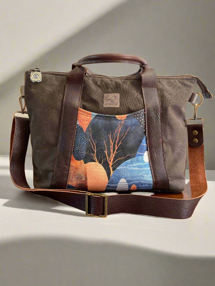 Oxbow crossbody/tote bag in waxed canvas exterior, with leather adjustable strap and handles. Two exterior front pockets, multiple inner pocket, fully lined. Chocolate brown waxed canvas, Land of Pebbles print on accent front and back pockets.