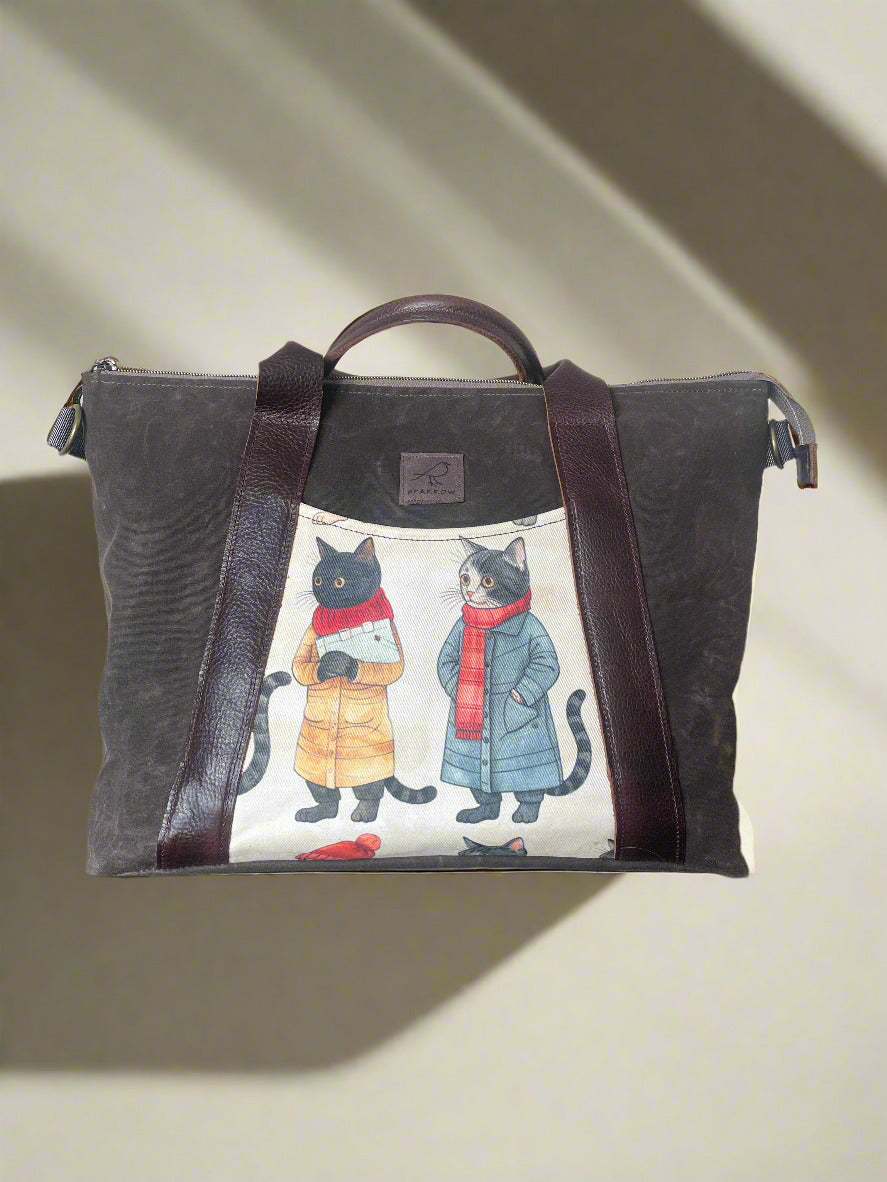 Large crossbody/handbag in dark brown waxed canvas. Cats wearing coats pattern on front and back accent pockets. Leather strap and handles.