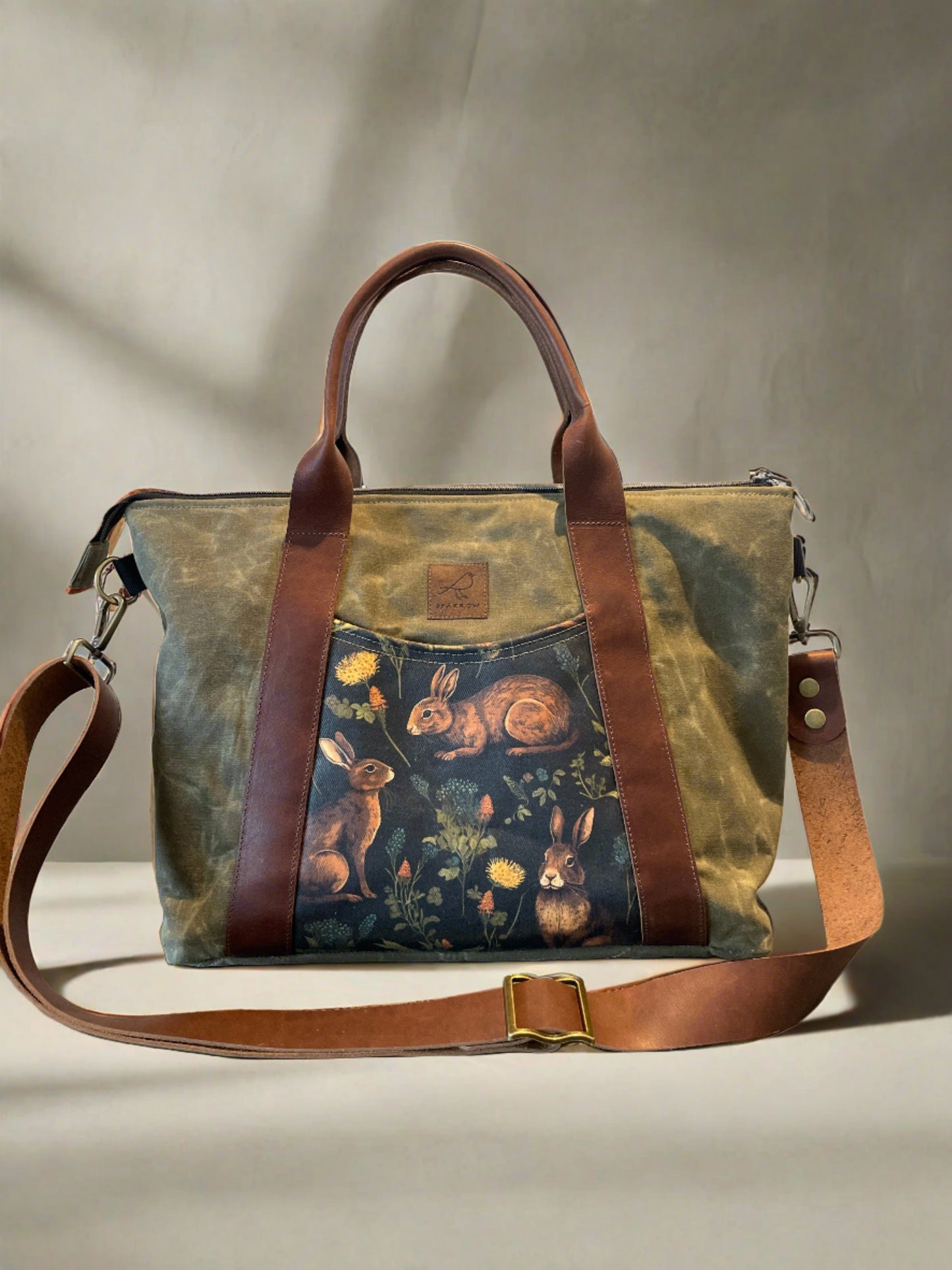 Oxbow crossbody/tote bag in waxed canvas exterior, with leather adjustable strap and handles. Two exterior front pockets, multiple inner pocket, fully lined. Olive green waxed canvas, rabbit print on accent front and back pockets.