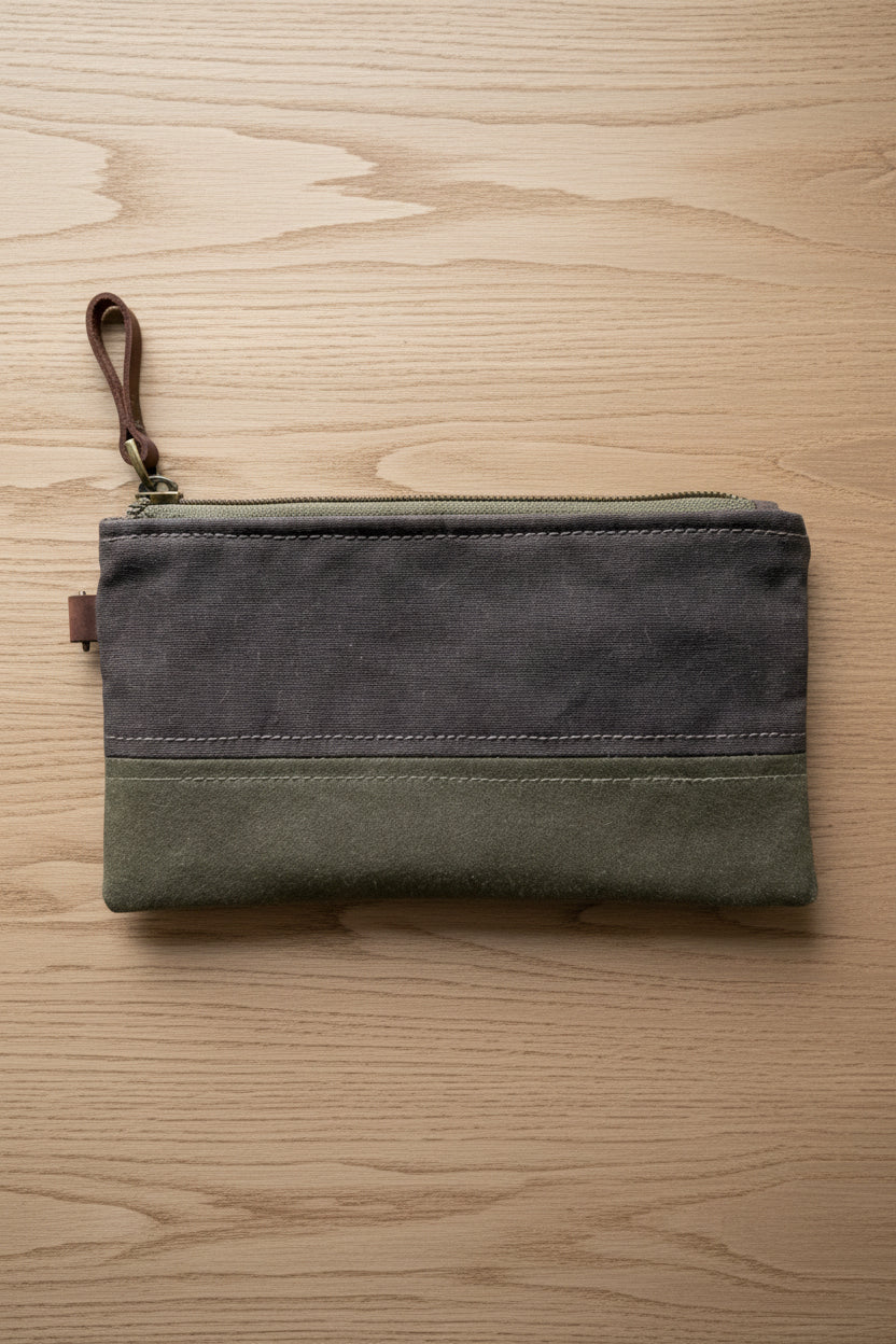 A rectangular waxed canvas zip pouch with a brown top half and an olive green bottom half, featuring a brown leather wrist strap and a gold-tone zipper, laid flat on a wooden background.