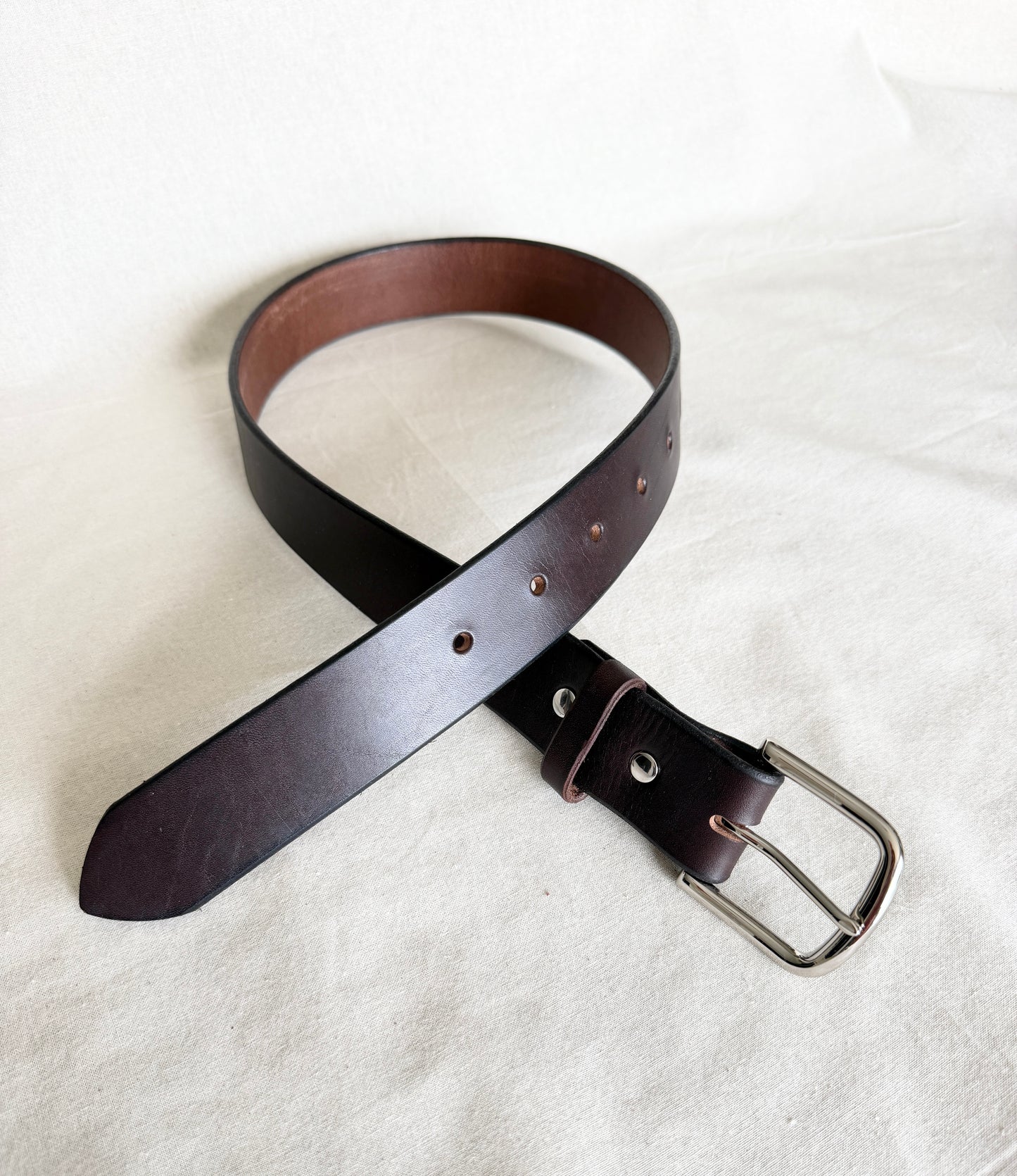 Dark brown leather belt with a silver buckle angled in a cream background to view both ends.