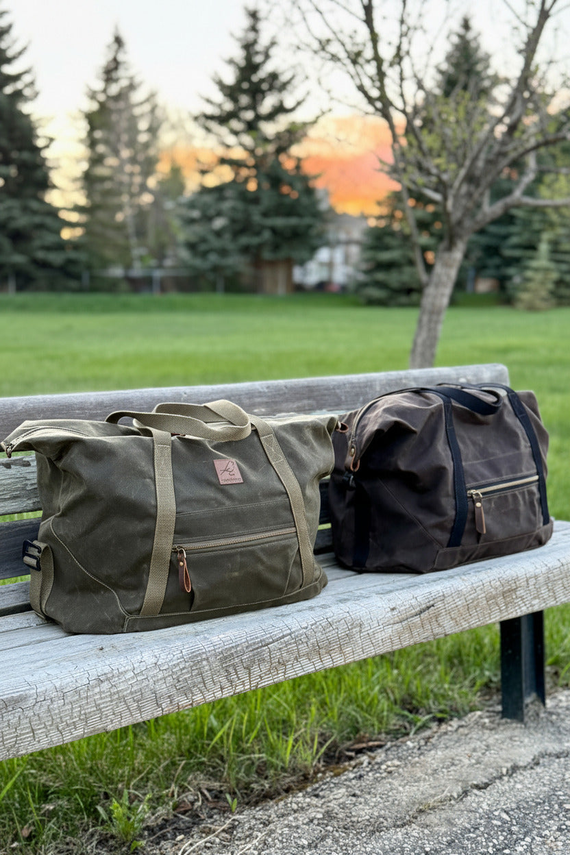 2 Weekender waxed canvas duffel bags on a bench - one in olive green, the other in chocolate brown and black straps