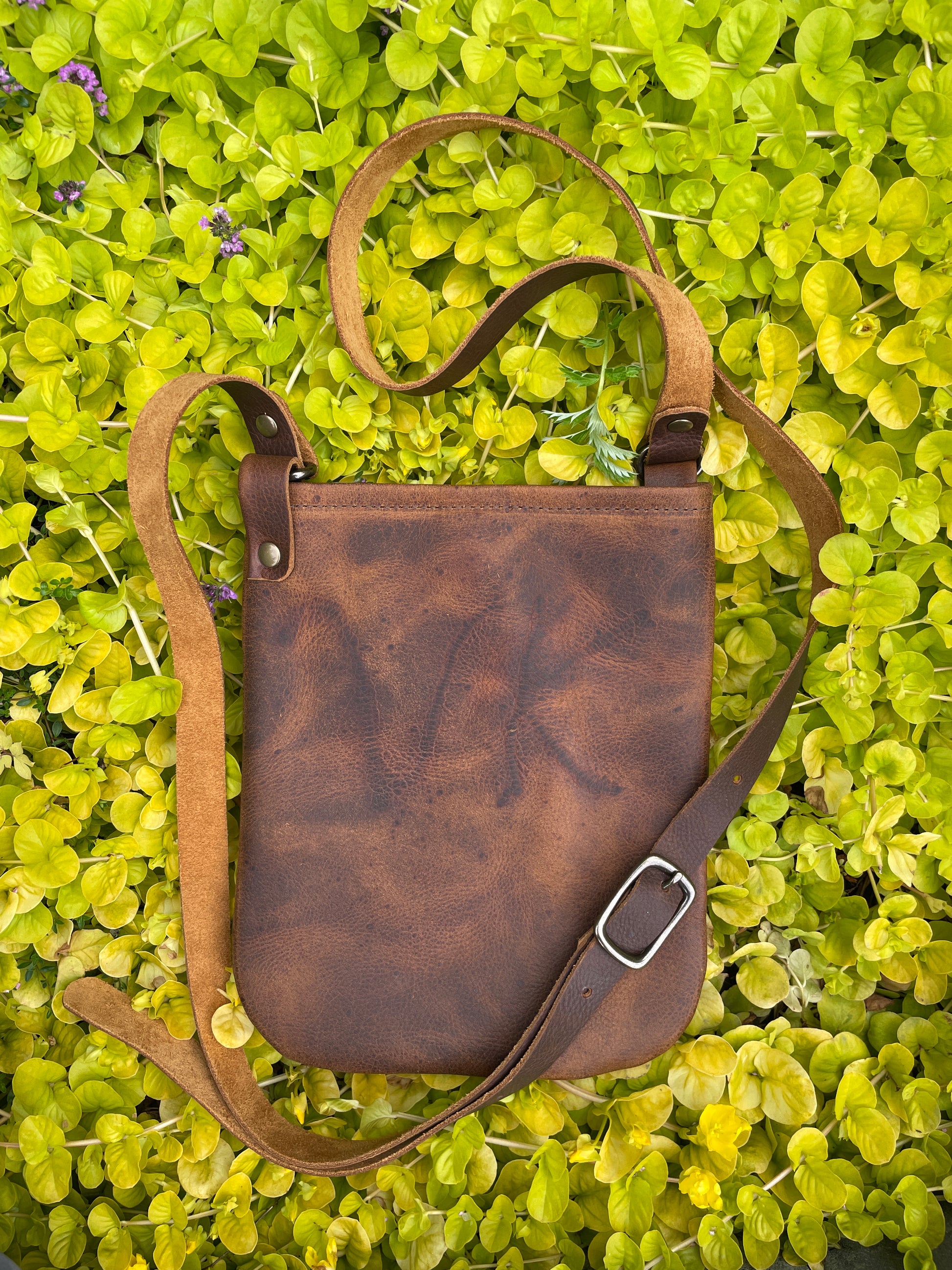 Brown leather bag with a strap on a green leafy background. Bag has initials VK