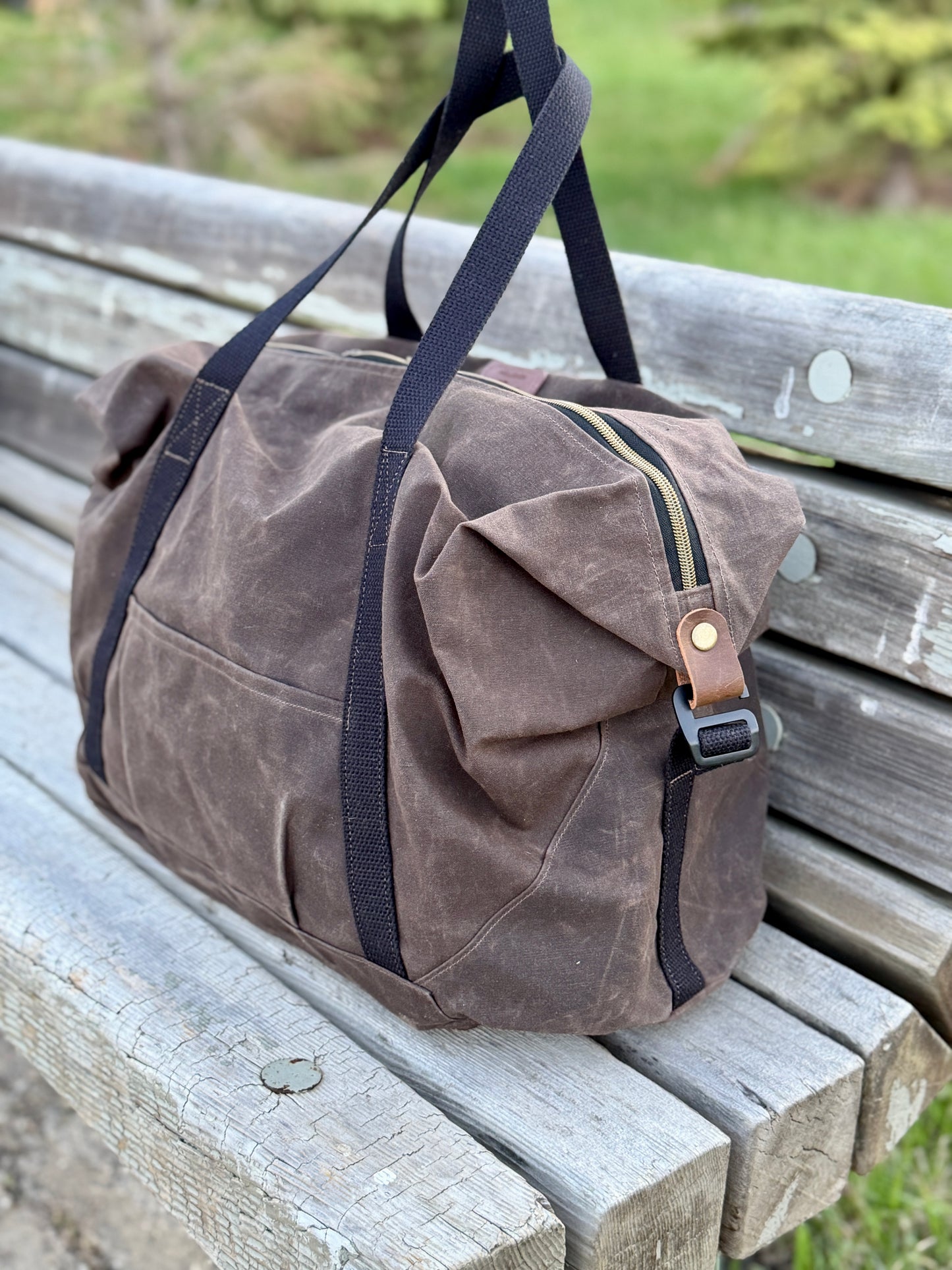 Brown waxed canvas duffel bag with black straps on a wooden bench outdoors.