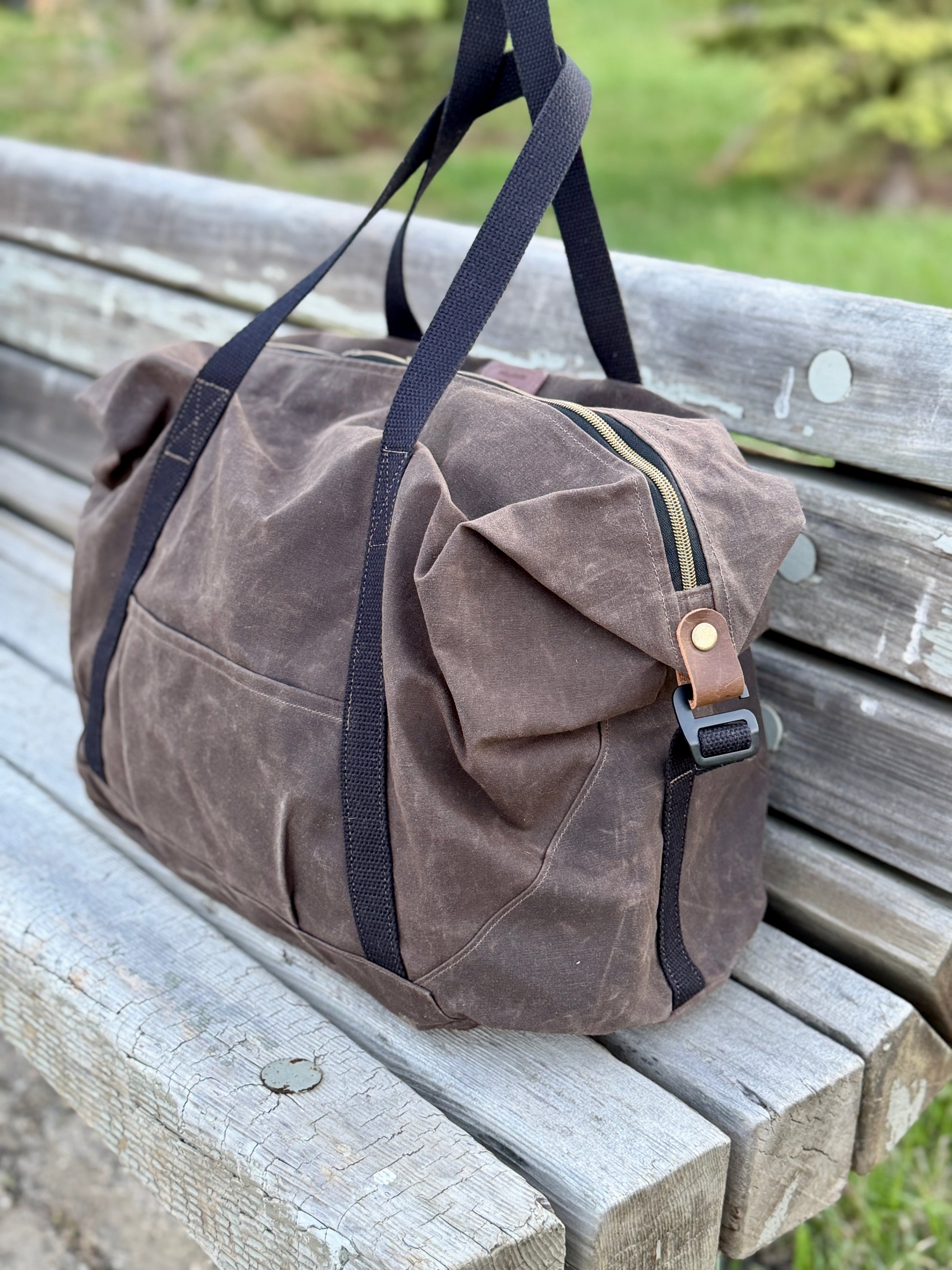 Brown waxed canvas duffel bag with black straps on a wooden bench outdoors.