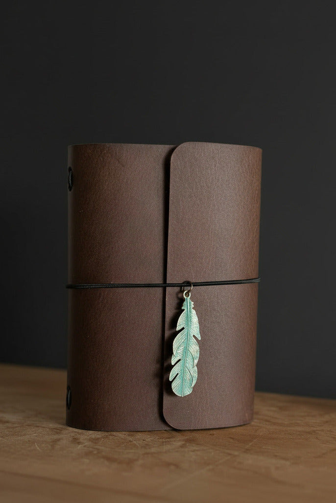 "Coffee" colored refillable leather traveler's journal with green metal feather embellishment on a desk, handcrafted in Calgary by Sparrow Handmade