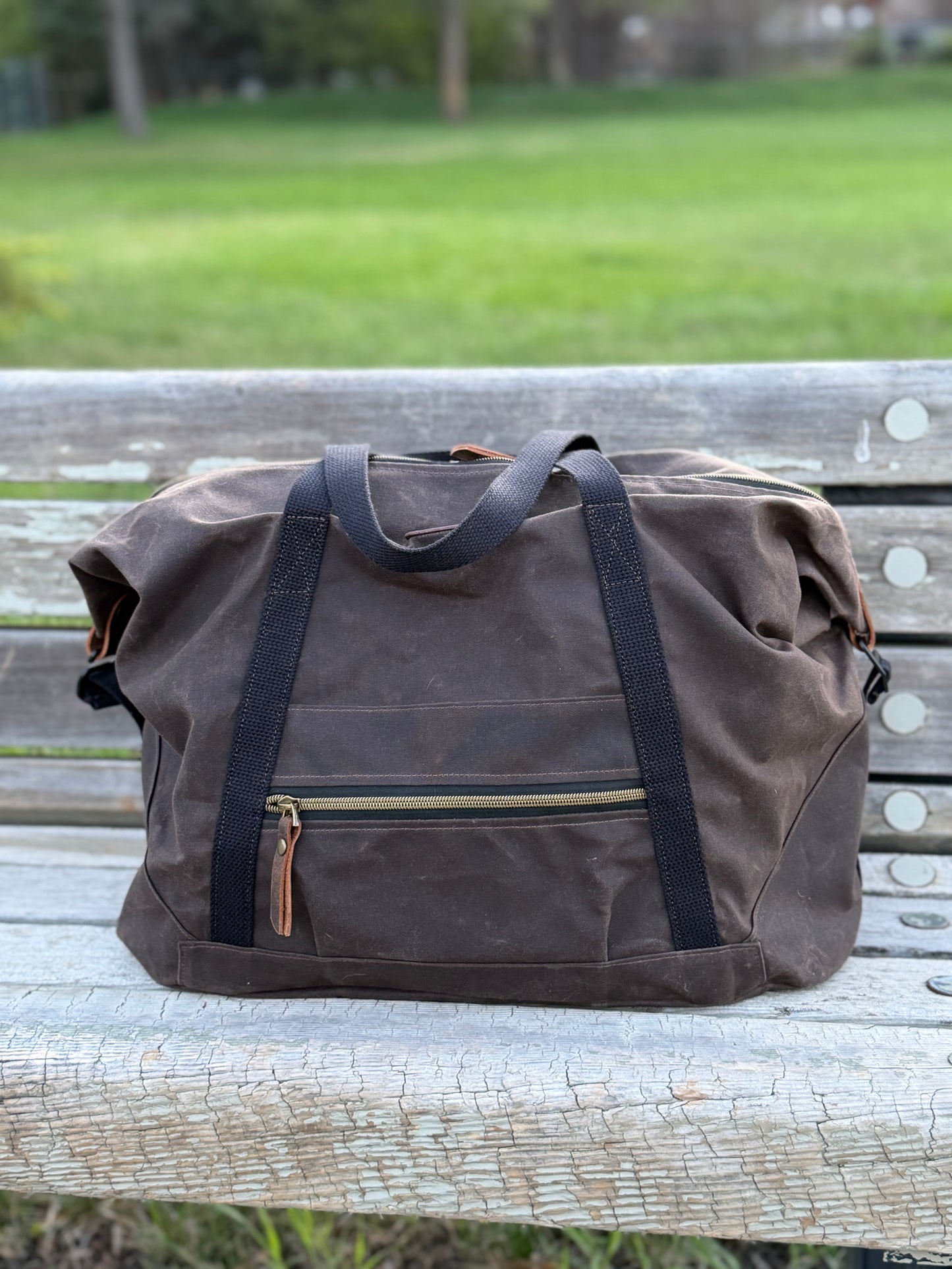 Brown weekender waxed canvas duffel bag with black straps on a wooden bench in a park.