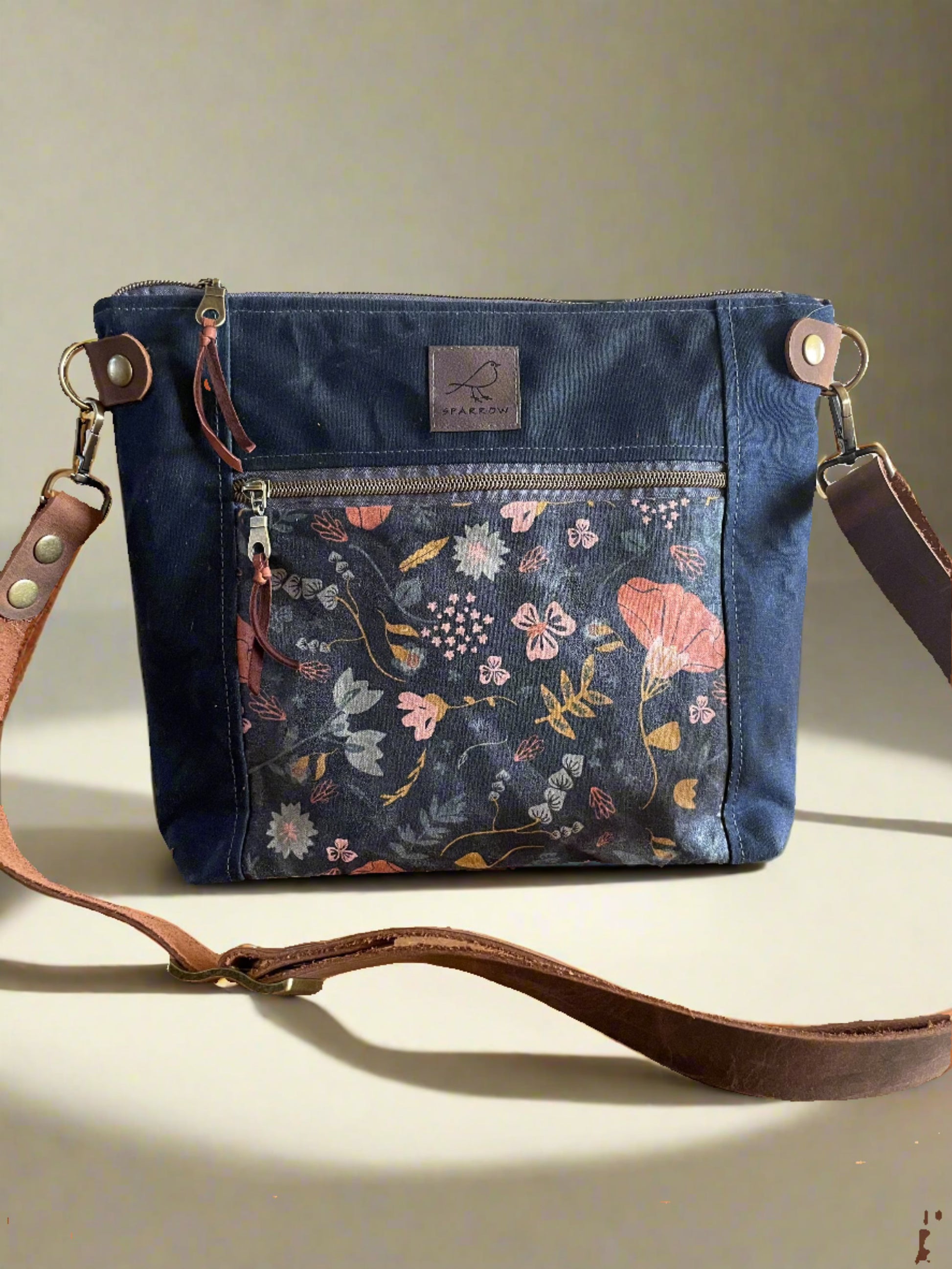 Cambridge crossbody bag in navy blue waxed canvas and coral floral front pocket accent print, with zippered front pocket and top closure. Adjustable leather strap. Handcrafted in Canada by Sparrow Handmade.