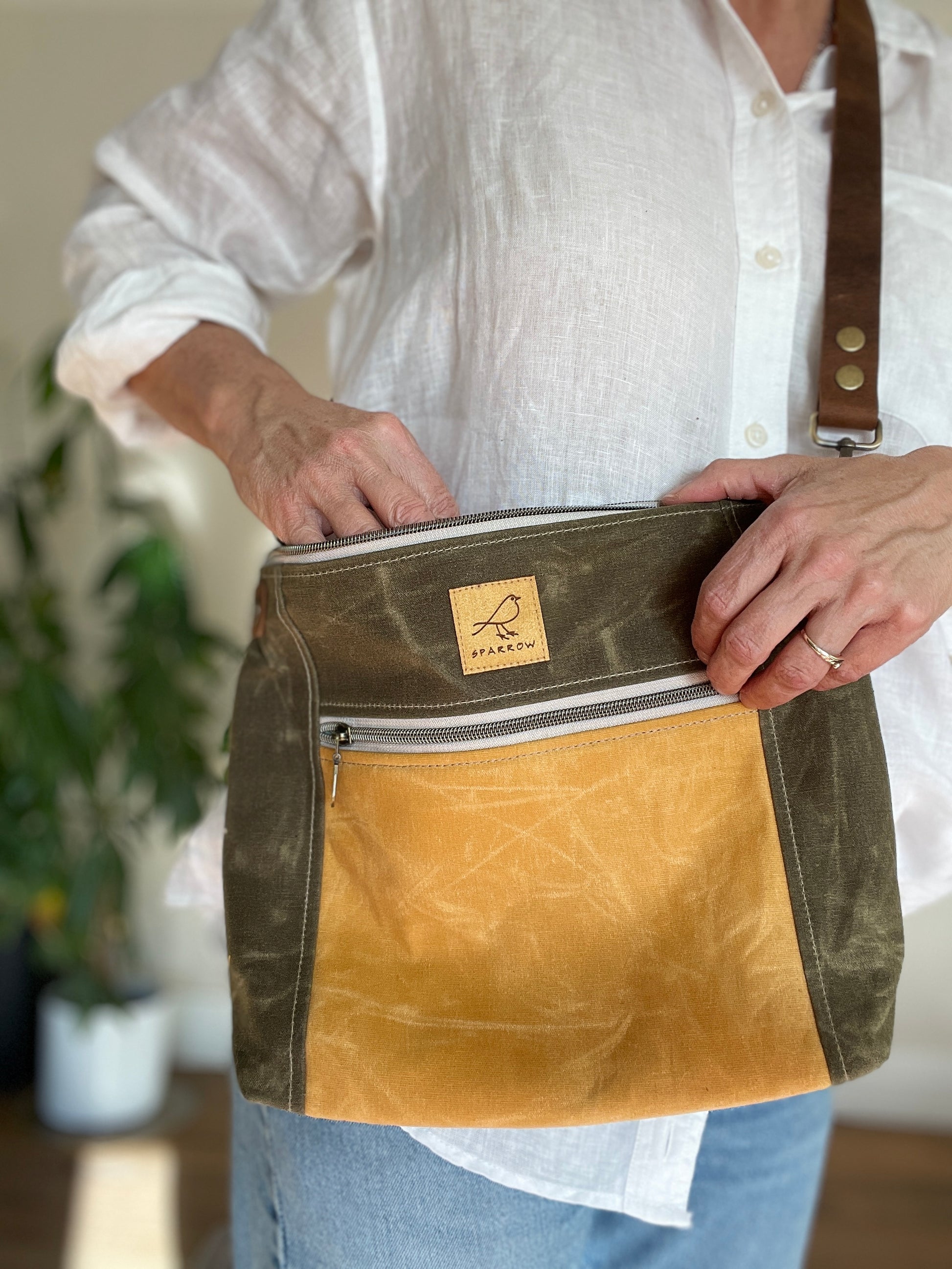 Cambridge crossbody bag in olive waxed canvas and ochre front pocket accent print, with zippered front pocket and top closure. Adjustable leather strap. Handcrafted in Canada by Sparrow Handmade. Worn by model in white shirt. 