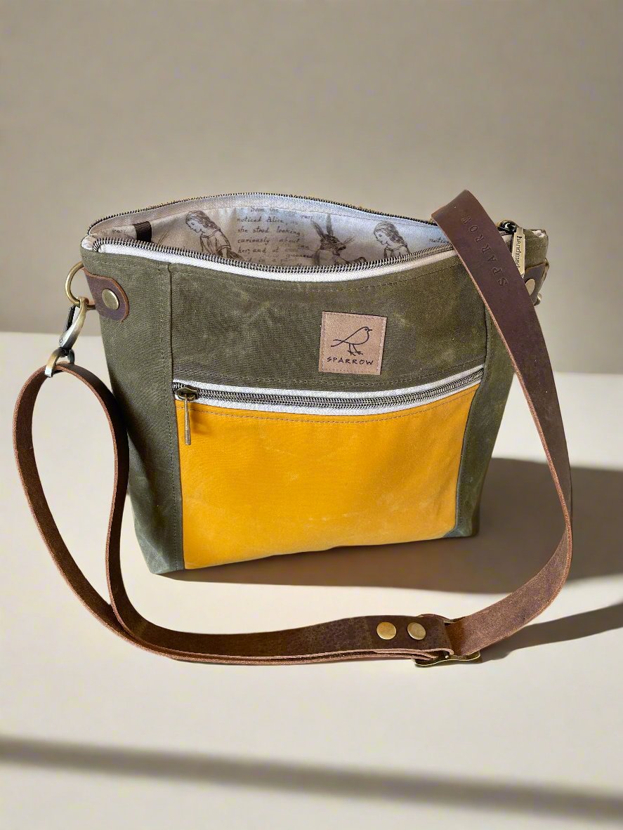 Cambridge crossbody bag in olivewaxed canvas and ochre pocket accent print, with zippered front pocket and top closure. Alice in Wonderland lining. Adjustable leather strap. Handcrafted in Canada by Sparrow Handmade.
