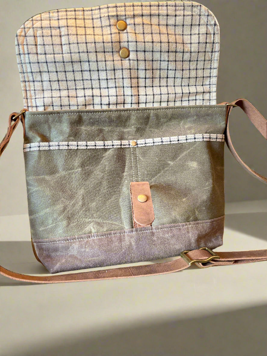 Shortie Messenger bag in olive green waxed canvas and a brown waxed canvas base. Brown leather 1.5 inch adjustable strap with a leather snap closure. Unisex bag. Bag is open showing a checkered lining. 