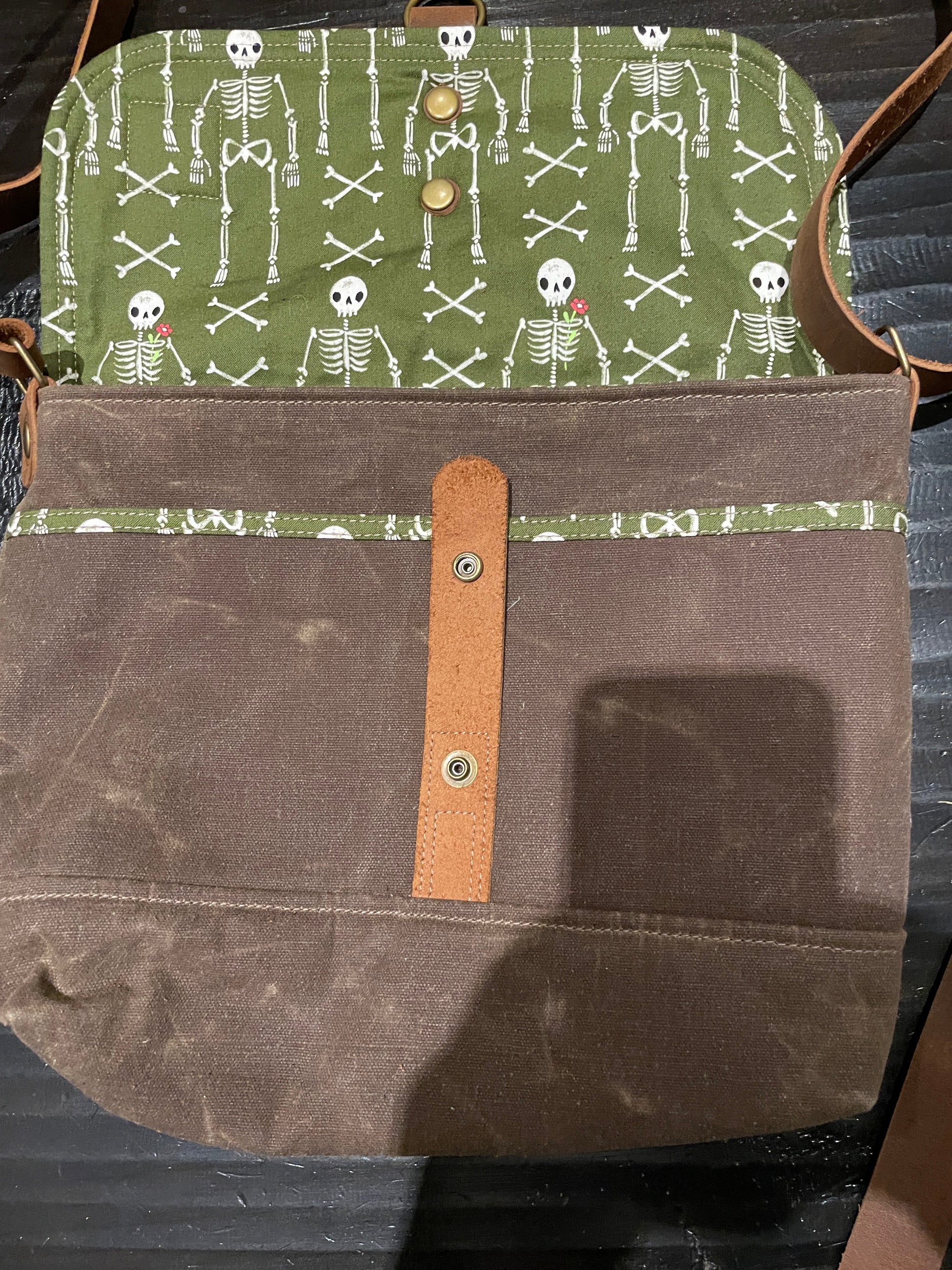 Shortie Messenger bag in brown waxed canvas and green and white patterned lining with skeletons. Brown leather 1.5 inch adjustable strap with a leather snap closure. Unisex bag. 