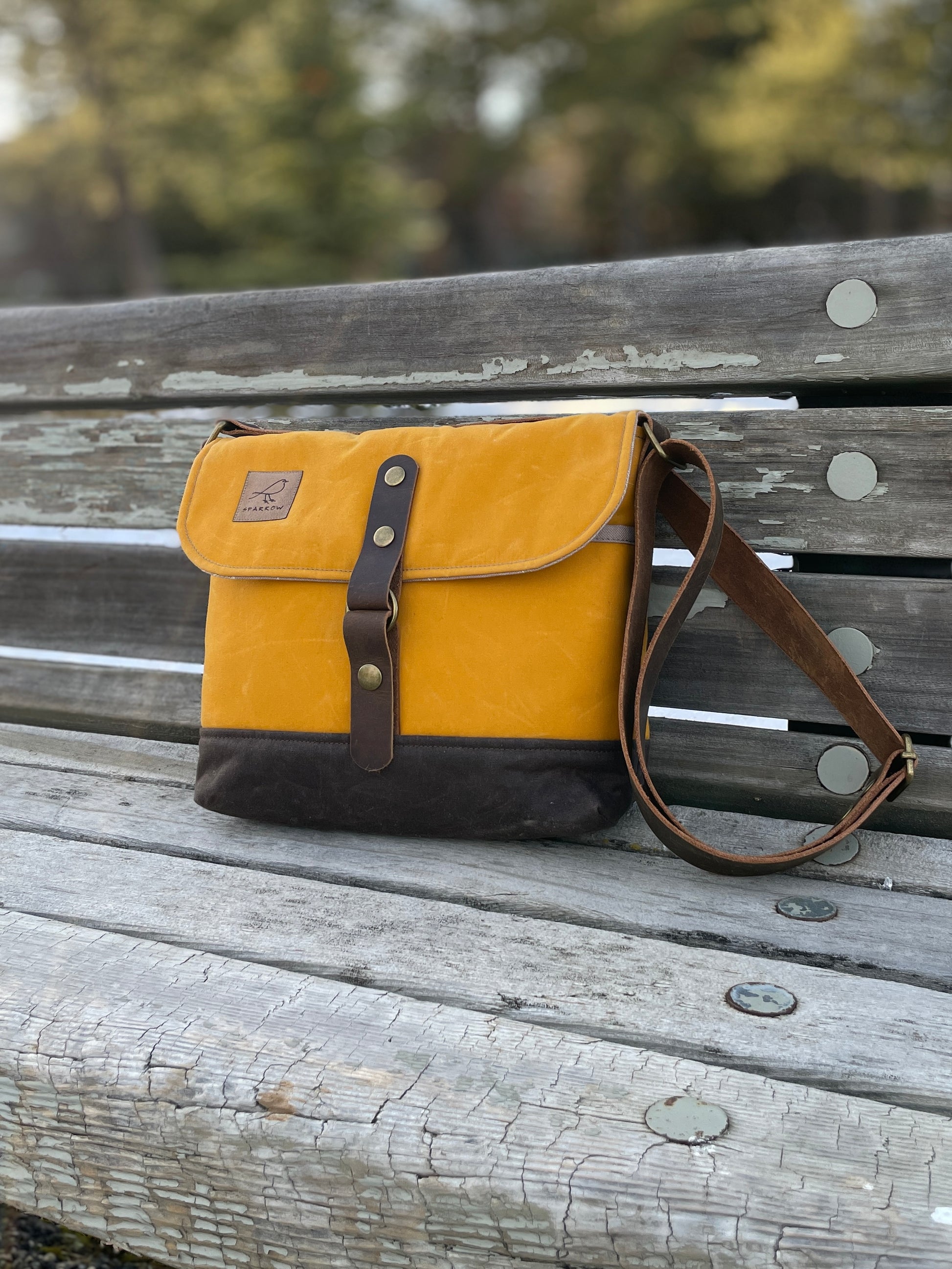 Waxed canvas messenger bag in ochre upper and chocolate brown base, with 1.5 inch wide adjustable full grain leather strap. Bag is sitting on a bench outside. 