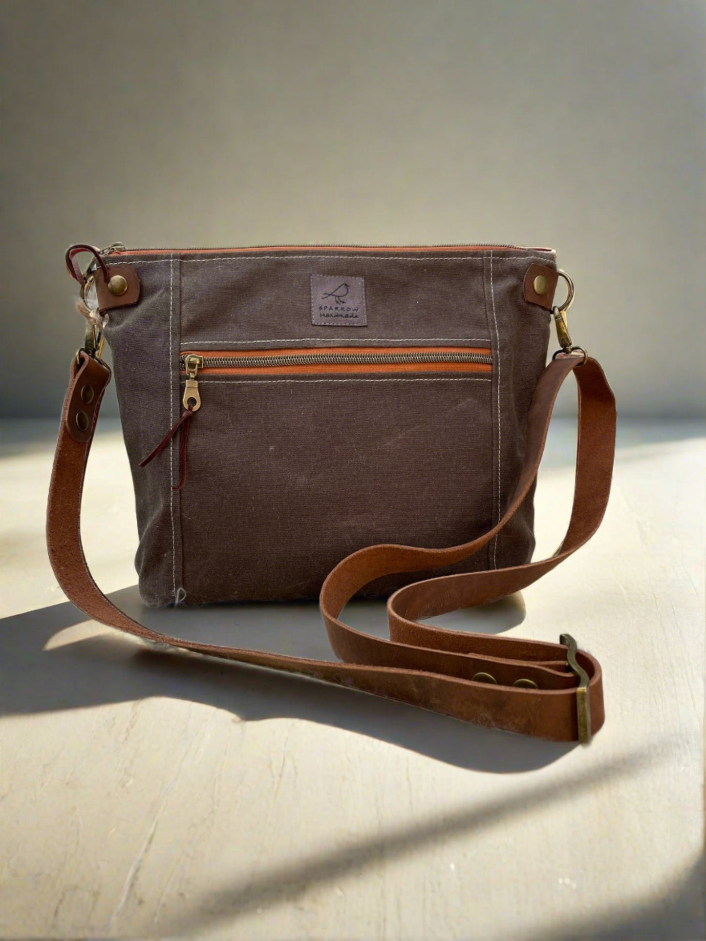 Brown waxed canvas crossbody bag, orange zipper with antique brass hardware. Adjustable leather strap, zippered main pocket, front zippered pocket, back slip pockets. Multiple interior pockets. 