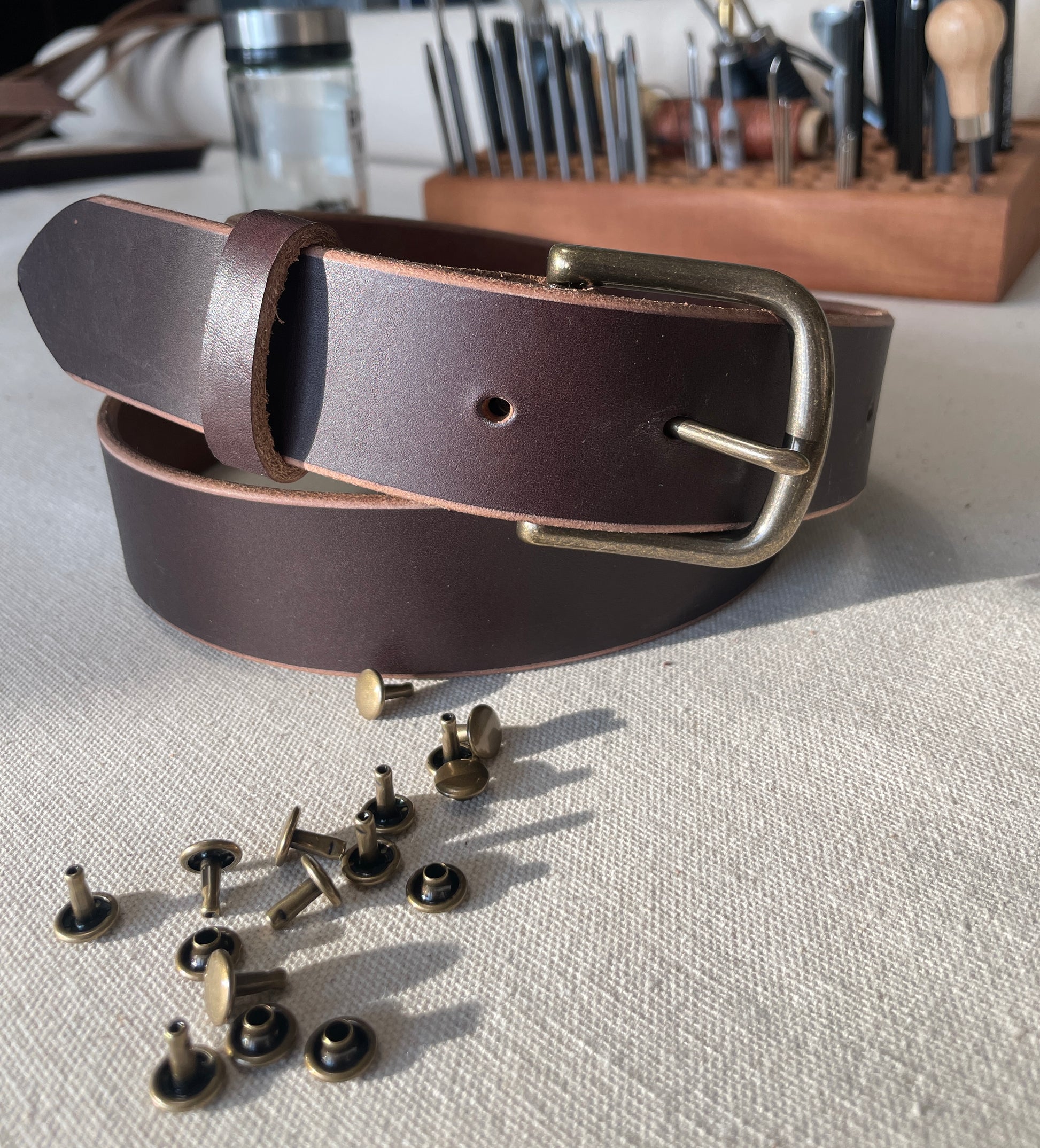 Custom handcrafted full-grain leather belt in Espresso brown with bronze hardware. Pictured with leatherworking tools. 