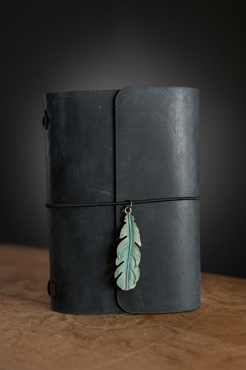 Slate black refillable leather traveler's journal with green metal feather embellishment on a desk, handcrafted in Calgary by Sparrow Handmade