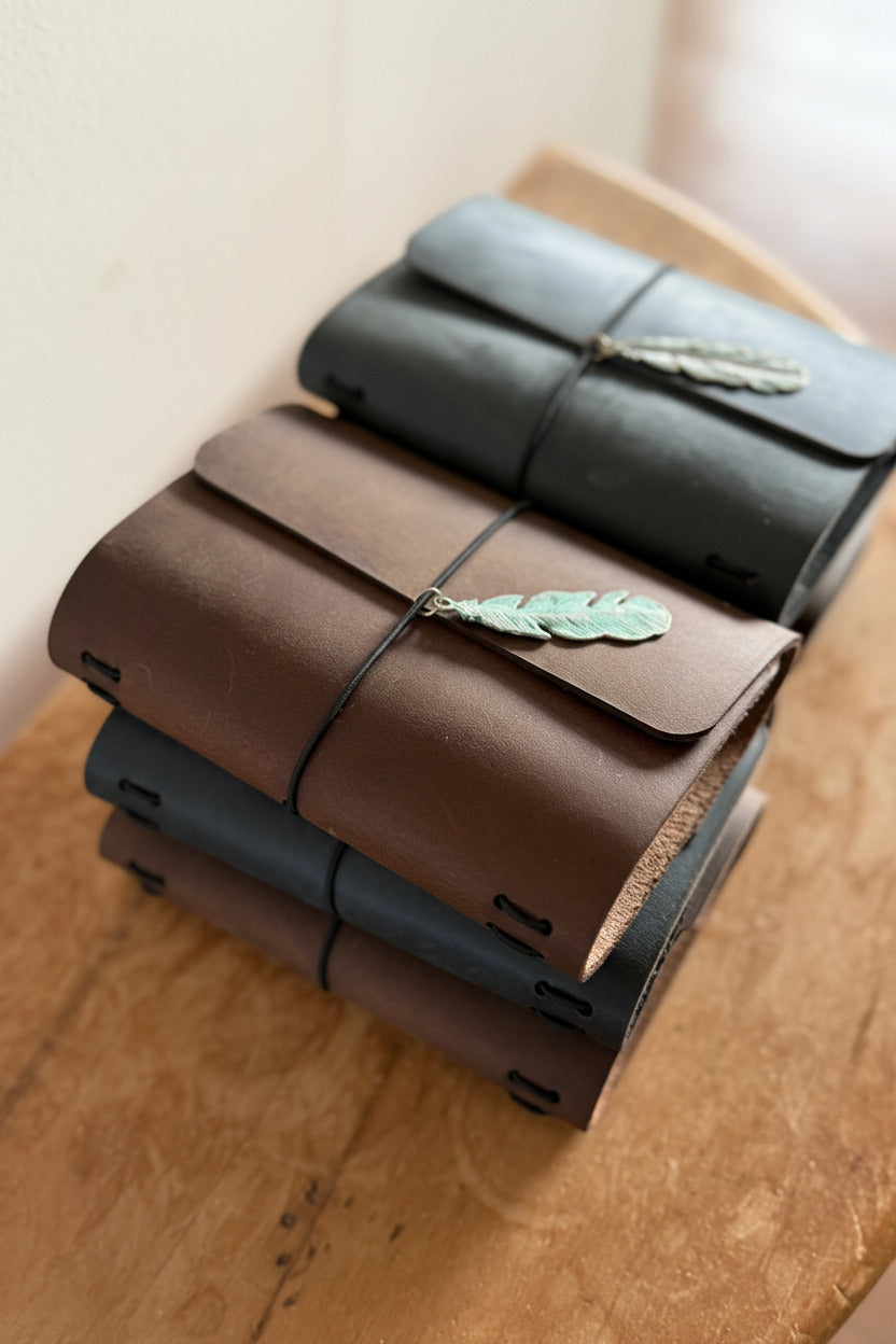 Stack of brown and black leather refillable traveler's journals on a desk. Journals are closed with a black elastic and embellished with a green metal feather. 