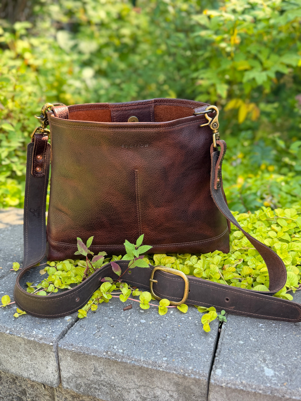 A dark brown leather shoulder bag with a matching adjustable strap and gold-tone buckle, resting on a stone ledge surrounded by green foliage.