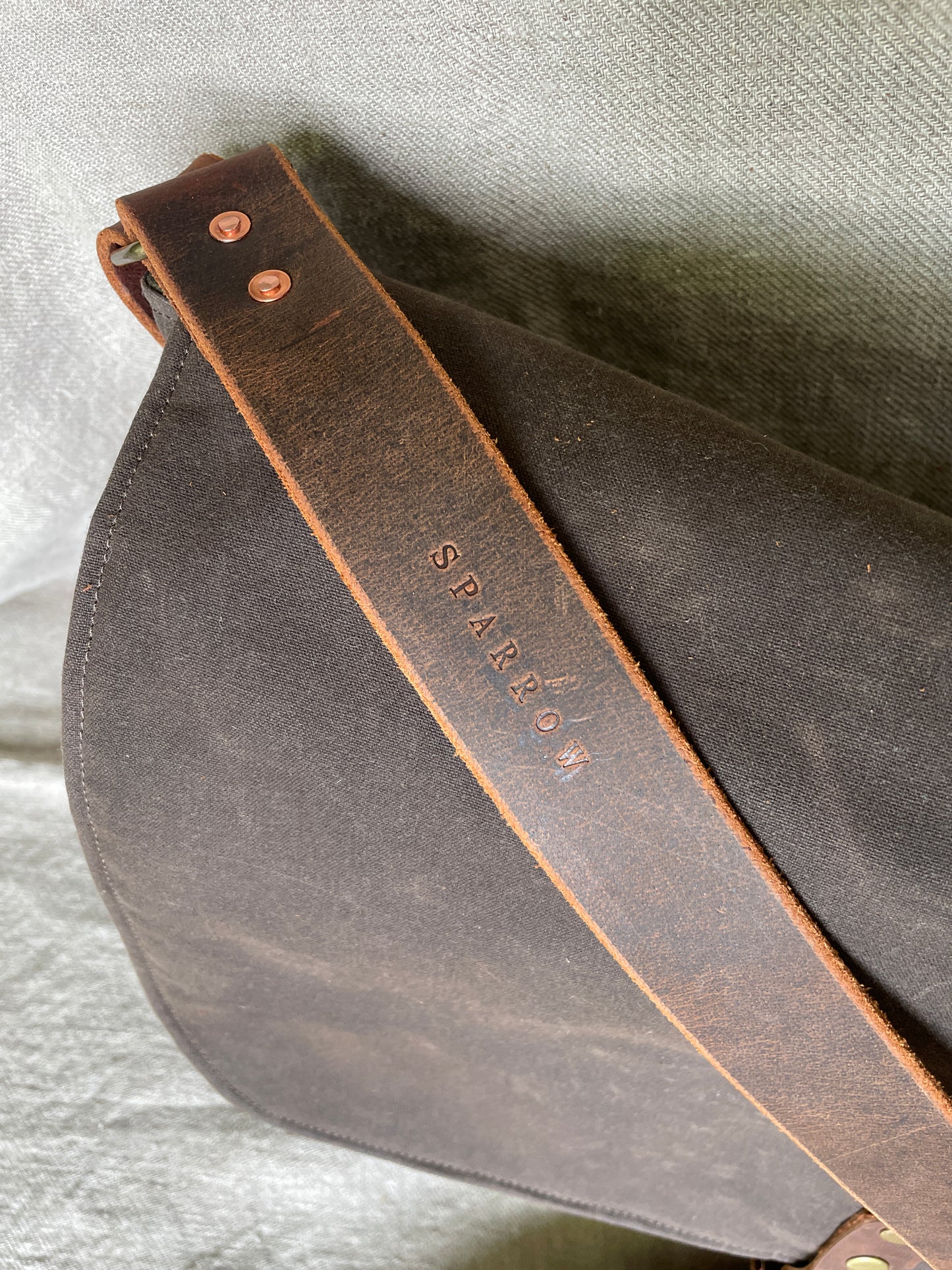 Detail photo of 1 1/2" leather strap with Sparrow logoand copper rivets on Attune crossbody bag in chocolate waxed canvas and olive green denim lining, for birthing stool. Leather strap, 4 leg pockets. 