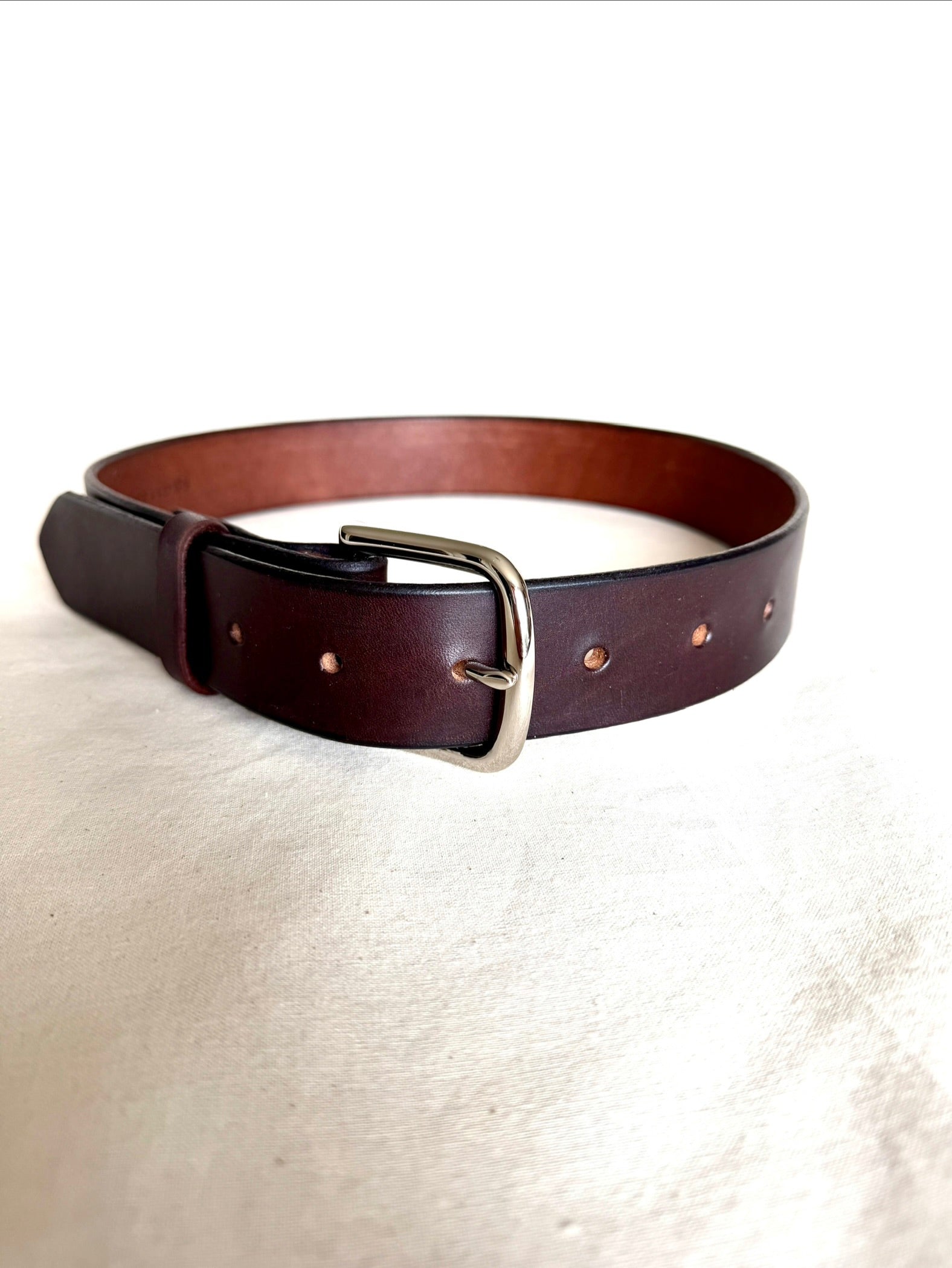 A dark brown leather belt with a silver-tone metal buckle, displayed on a textured off-white fabric background.