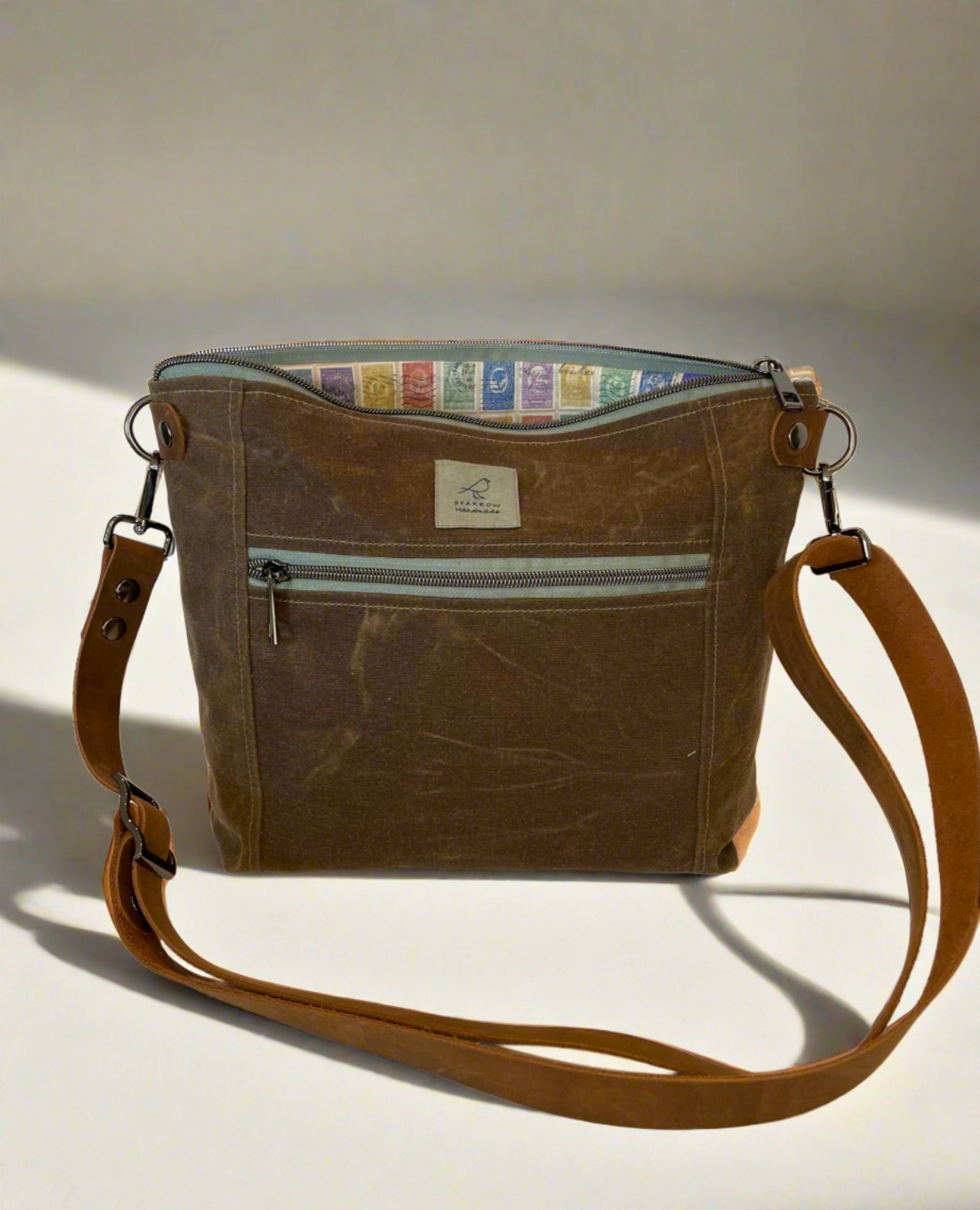 Cambridge crossbody bag in brown waxed canvas with zippered front pocket and top closure. Colorful stamps lining, mint green zipper and gunmetal hardware. Adjustable leather strap. Handcrafted in Canada by Sparrow Handmade.